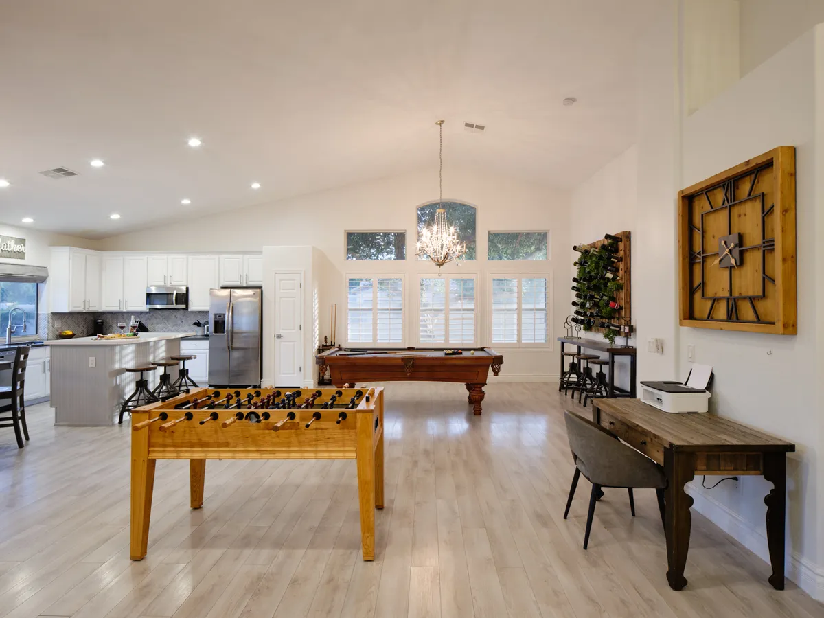 A vibrant, open interior featuring a foosball table and pool table, with elegant lighting from a chandelier and natural light streaming through large windows. A workspace with a desk is visible, blending gaming and productivity in one communal area.