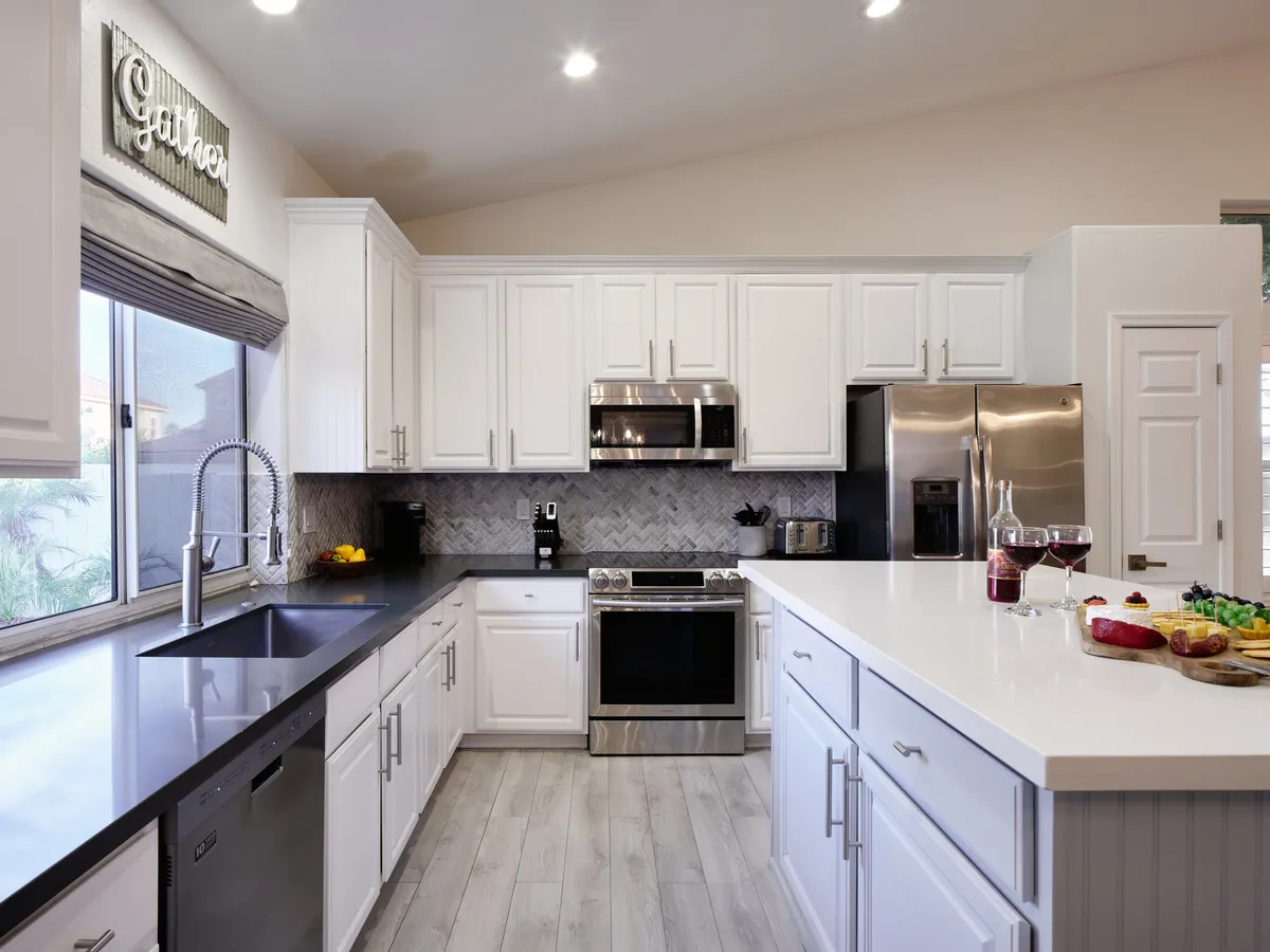 The image features a bright, airy kitchen and dining area with a gathering table set with food and drinks. Stainless steel appliances and white cabinetry create a modern, clean look, while natural light streams in through a window, enhancing the inviting mood.
