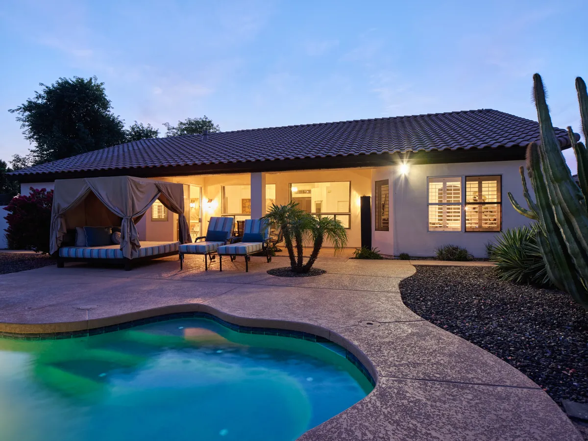 A stylish evening pool scene with an outdoor seating area under a canopy lounge, surrounded by a landscaped garden. Warm lighting suggests a comfortable, inviting ambiance perfect for stargazing or evening gatherings.