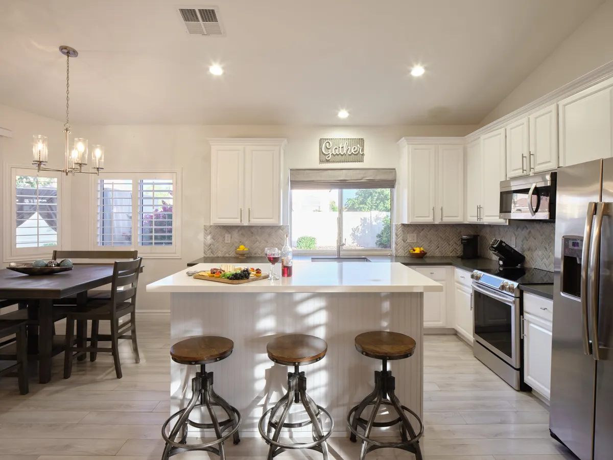 A cozy kitchen scene with an island counter and comfortable seating, complemented by a dining table set near a window offering an inviting view. The space is bathed in soft, warm light, emphasizing the welcoming feeling for both meal prep and dining.