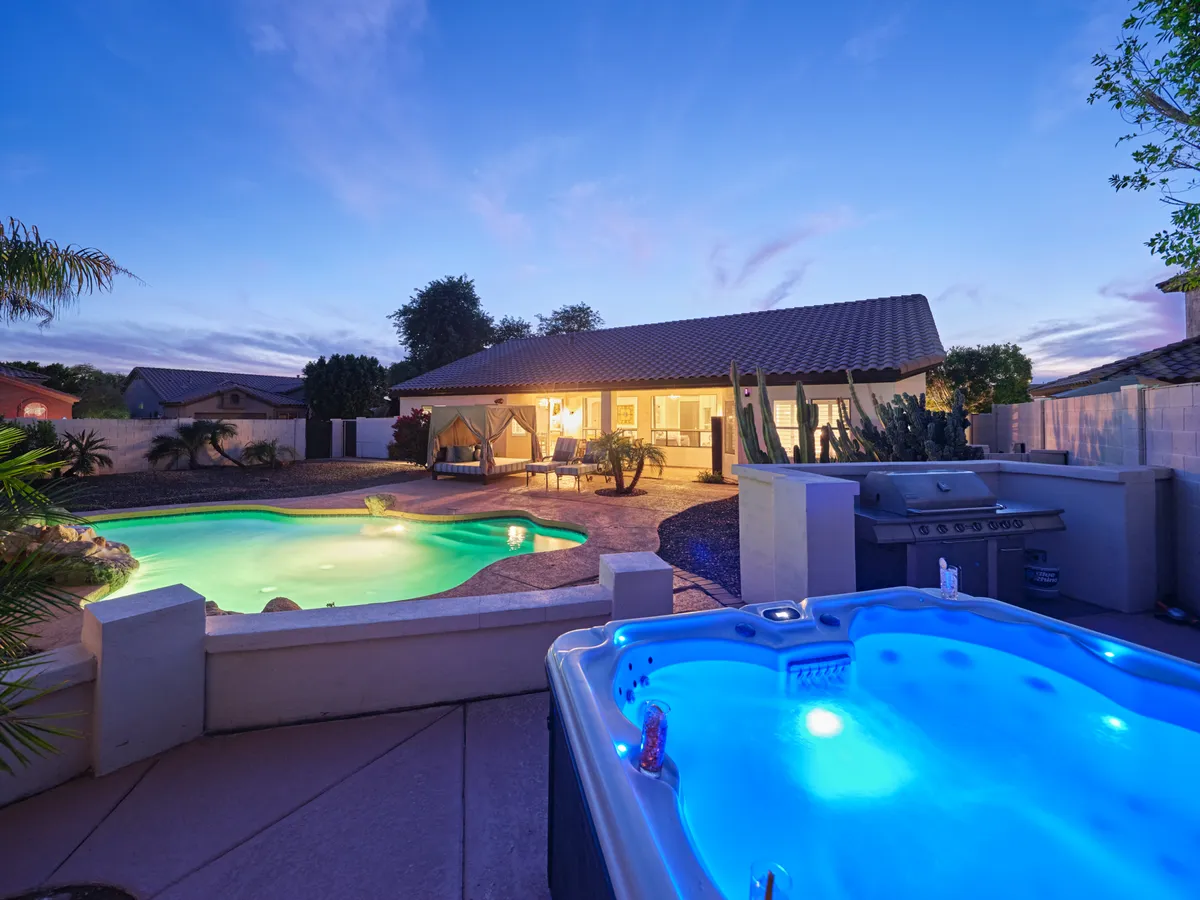 The image showcases a stunning Scottsdale backyard oasis featuring a sparkling swimming pool, a bubbling hot tub, stylish outdoor seating, and lush desert-inspired landscaping. The tranquil setting is perfect for relaxing, entertaining, or enjoying vibrant sunsets with friends and family.