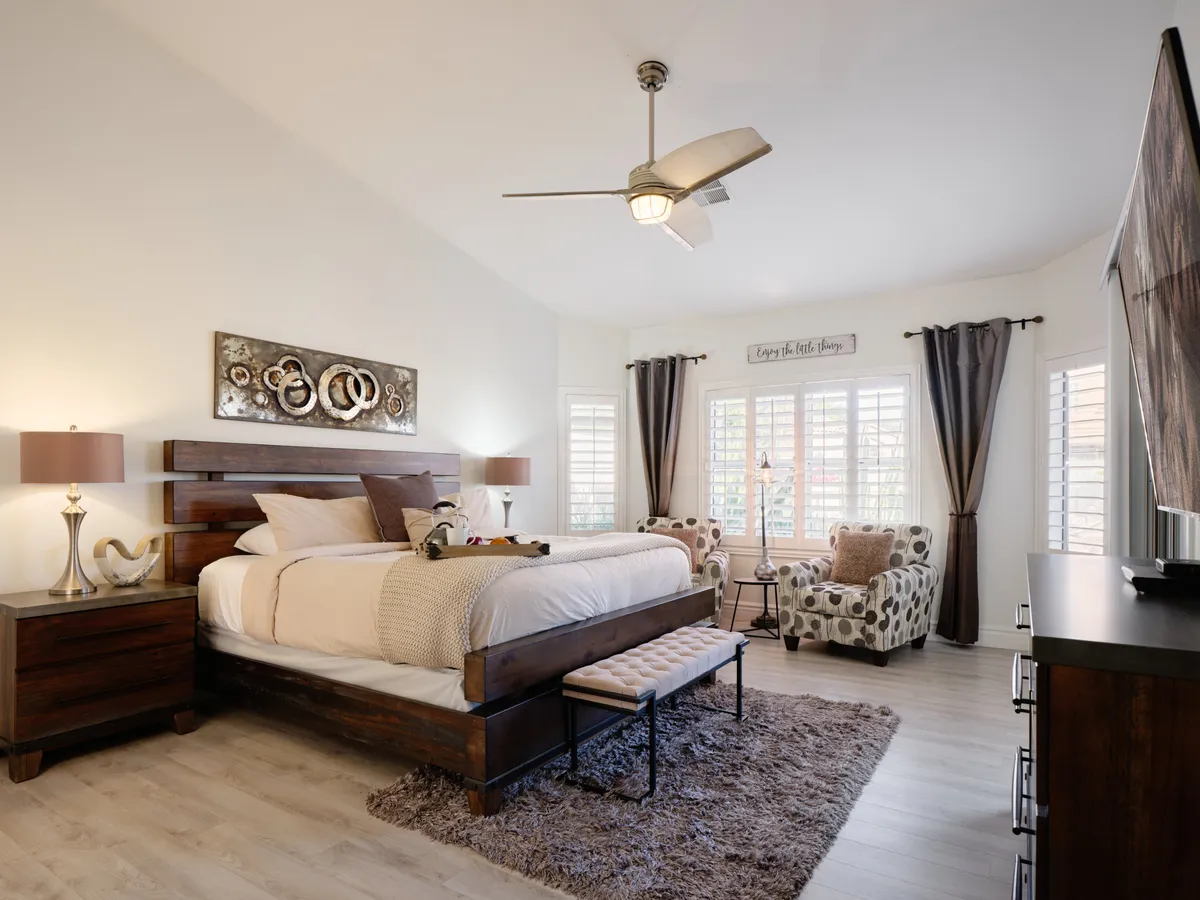 A bright, spacious bedroom featuring a king-sized bed adorned with decorative pillows, stylish wall art above the headboard, cozy armchairs, and matching nightstands. The room is filled with natural light, creating a serene and comfortable atmosphere—ideal for rest and relaxation.