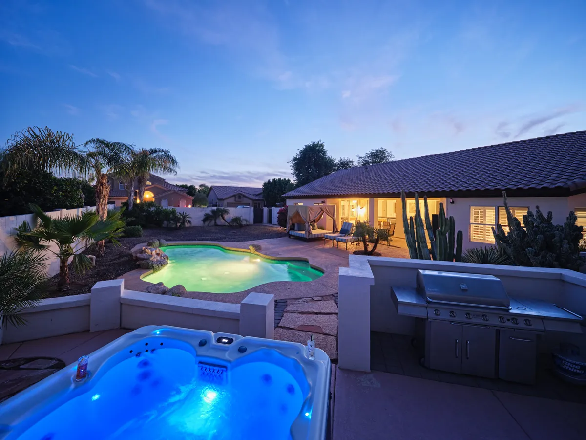 A serene backyard oasis with a jacuzzi, swimming pool, comfortable outdoor seating, a grill, and desert landscaping dotted with cacti. The warm lighting suggests a relaxing evening perfect for post-golf unwinding.
