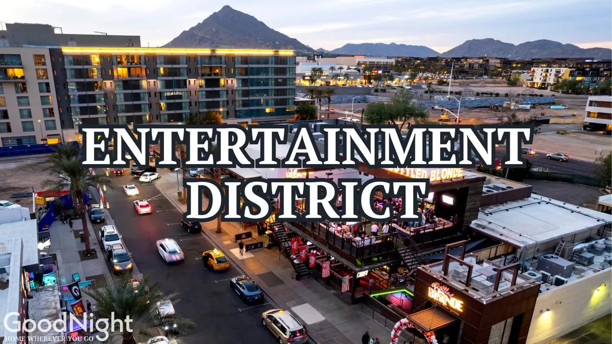 This exterior scene captures the vibrant Scottsdale Entertainment District, with visible signage, bustling outdoor dining areas, parked cars, and a mountain backdrop. There is a lively, energetic atmosphere that hints at nightlife, social activity, and the appeal of city exploration after sunset.