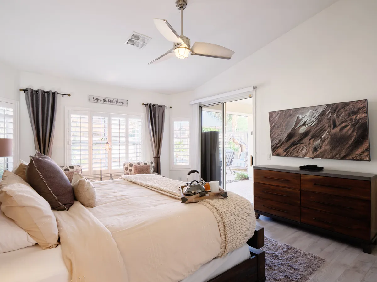 A king bedroom with a sleek bed dressed in decorative pillows, a wall-mounted TV opposite the bed, nightstand with tray, and sunlight streaming in from a generous window. The scene is inviting and perfect for restful moments or unwinding with entertainment.